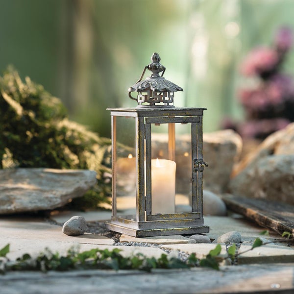 Rustikale Metalllaterne mit Glasscheiben und brennender Kerze, platziert auf Steinplatten in einer Gartenlandschaft.