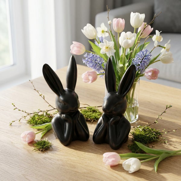Zwei Osterhasen aus Keramik als Tischdekoration neben einem Blumenstrauß mit Frühlingsblumen auf einem Holztisch.