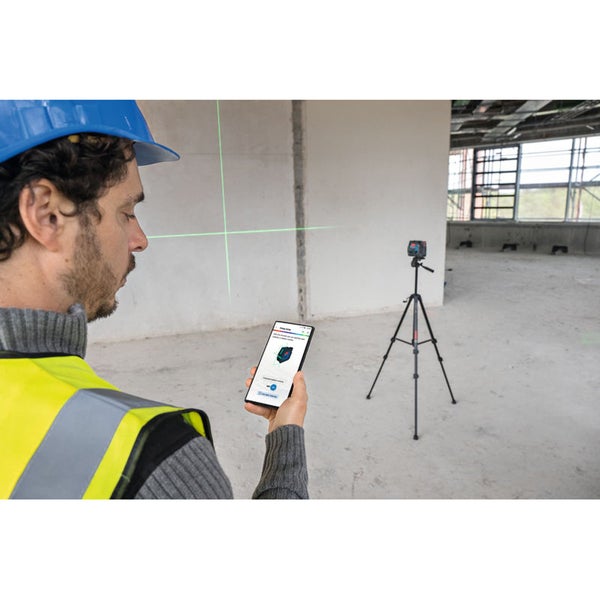 Ein Bauarbeiter mit Schutzhelm und Warnweste bedient eine Nivelliergerät-App auf seinem Smartphone, wobei ein Kreuzlinienlaser grüne Linien an die Wand projiziert. Ein Laser-Nivelliergerät steht auf einem Stativ im Hintergrund.
