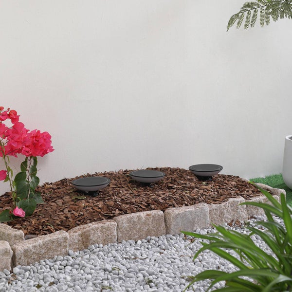 Drei runde Solar-Gartenleuchten in einem mit Rindenmulch gefüllten Beet, eingefasst von hellen Natursteinen und weißen Kieseln.