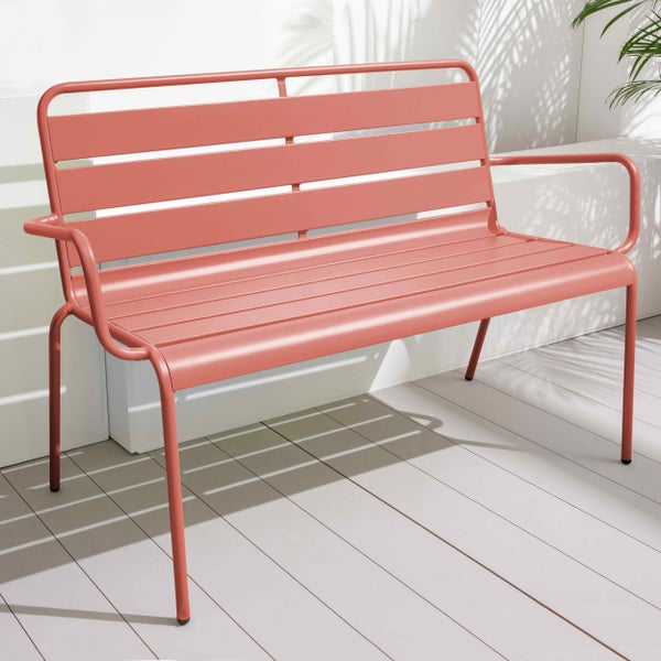 Moderne lachsrosa Gartenbank aus Metall mit horizontalen Latten auf einer hellen Holzterrasse mit Schatten von Palmenblättern.