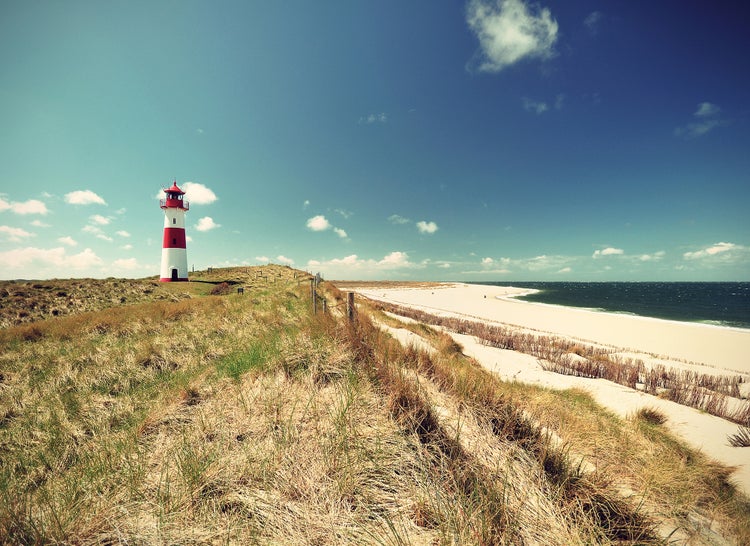 Wandbild mit Leuchtturm in einer Dünenlandschaft am Sandstrand mit Blick auf das Meer.