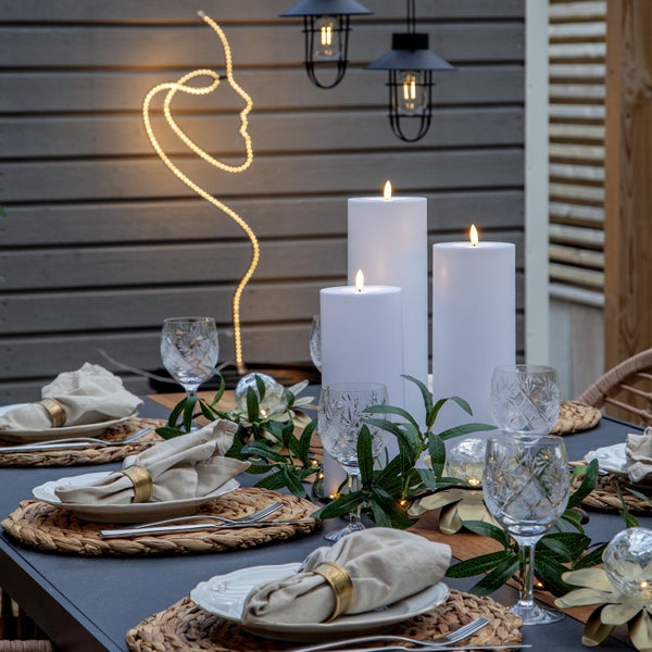 Gedeckter Esstisch mit Kerzen, Gläsern, Geschirr, Servietten und dekorativer Beleuchtung für ein stimmungsvolles Ambiente im Freien.