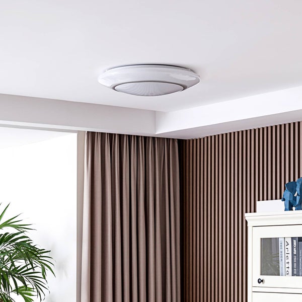 Moderne LED Deckenleuchte mit Lautsprecher an weißer Decke in einem Raum mit braunen Vorhängen und Holzlamellenwand.