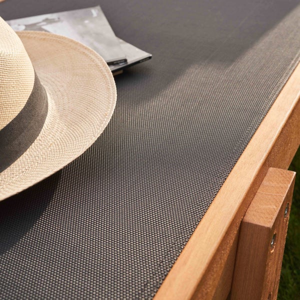 Detailaufnahme einer Gartenliege mit Holzgestell, dunklem Netzgewebe, einem Strohhut und einer Broschüre.