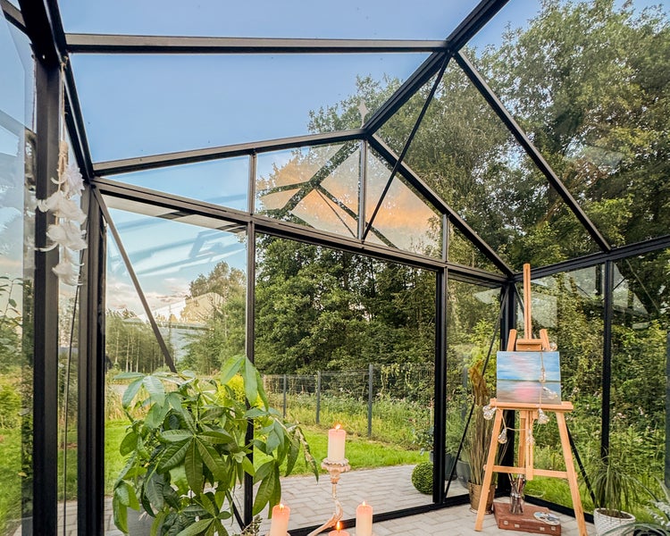 Modernes Gewächshaus mit schwarzem Metallrahmen, Glaswänden, Zimmerpflanzen, Kerzen und einer Staffelei in heller Atmosphäre.