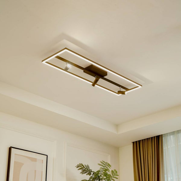 Moderne goldfarbene LED-Deckenleuchte mit rechteckigem Rahmen und drei Strahlern in einem hellen, organisierten Wohnraum mit Pflanzen und Vorhängen.