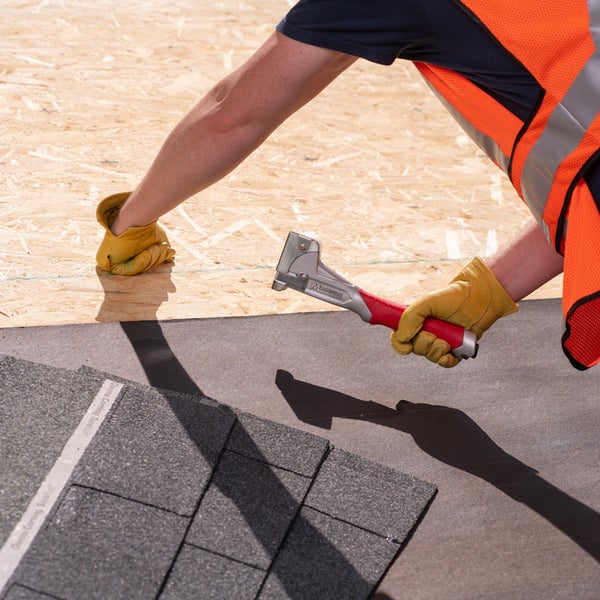 Bauarbeiter nutzt einen Hammertacker zum Befestigen von Dachschindeln auf OSB-Platten, trägt eine Warnweste und Arbeitshandschuhe.
