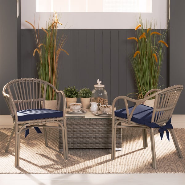 Garten-Lounge-Set aus Rattan mit zwei Sesseln, blauen Sitzkissen und einem Glastisch, dekoriert mit Pflanzen und Geschirr auf einem Teppich.