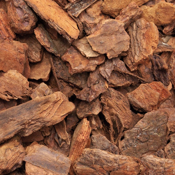 Galamio Pinienborke wird in einem hellen Garten um eine Lavendelpflanze verteilt. Ein Sack mit Galamio Logo und Gartenwerkzeug sind sichtbar.