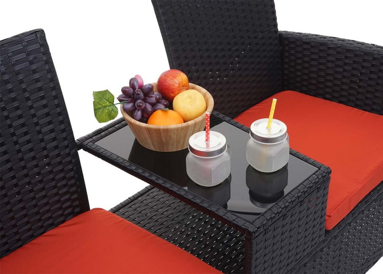 Gartenbank aus Polyrattan mit integriertem Glastisch zwischen zwei Sitzen, rote Sitzkissen, Obstschale und Trinkgläser auf dem Tisch.