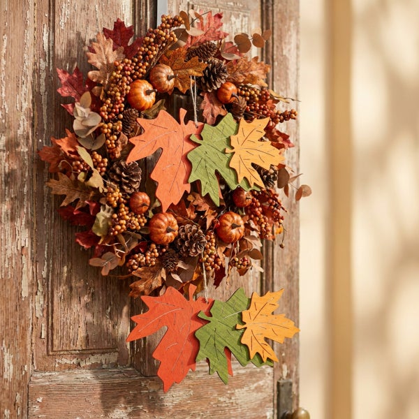 Herbstlicher Türkranz aus Kunstlaub, Tannenzapfen und kleinen Kürbissen mit hängenden Dekoblättern aus Holz an einer rustikalen Holztür.