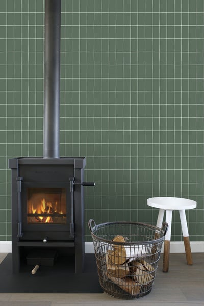 Kaminofen mit Holzkorb und Hocker vor einer Wand mit quadratischem Muster.