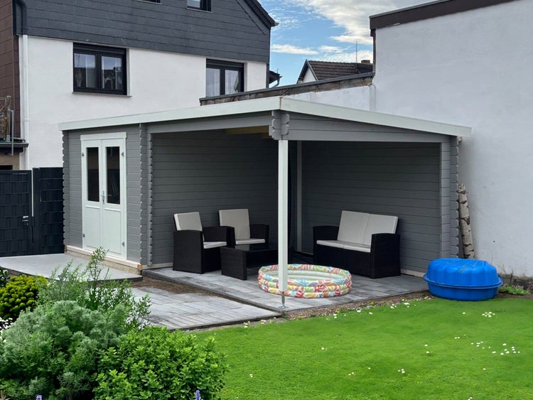 Gartenhaus aus Holz in Grau mit Pultdach, Doppeltür mit Lichtausschnitten und integriertem Terrassenbereich mit Gartenmöbeln auf einem Rasenstück.