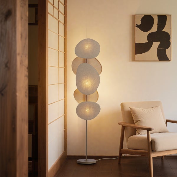 Moderne Stehleuchte mit fünf organisch geformten Textilschirmen in einem hellen Wohnzimmer neben einem Holzsessel.