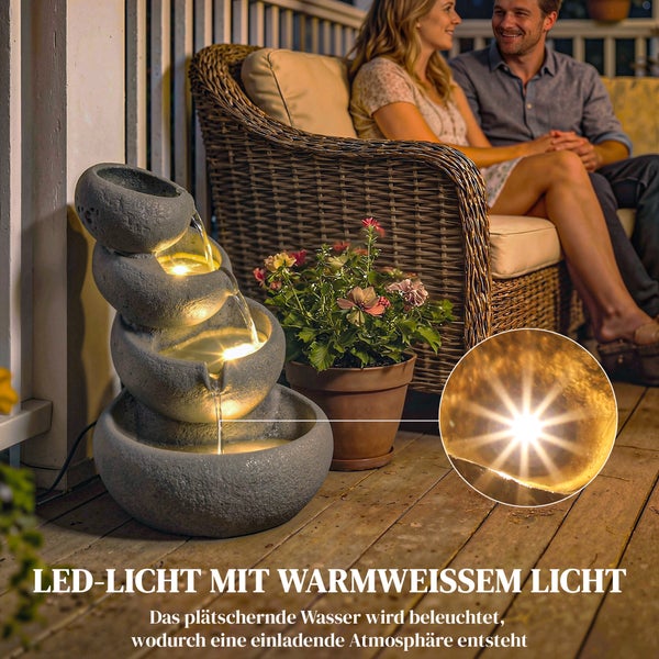Etagenbrunnen in Steinoptik mit vier Kaskadenschalen und LED-Beleuchtung in einer Gartenlandschaft mit Blumen.