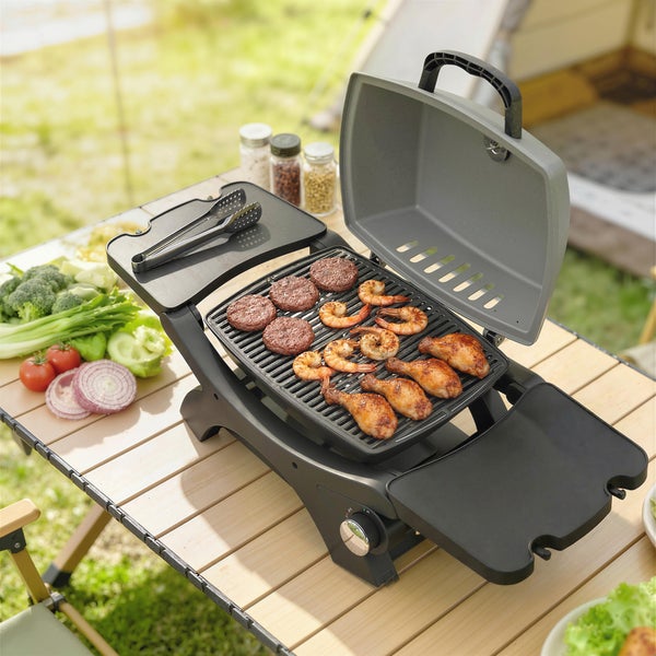 Mobiler Tischgrill auf einem Holztisch beim Camping mit gegrillten Burgern, Garnelen, Hähnchenkeulen und frischem Gemüse.