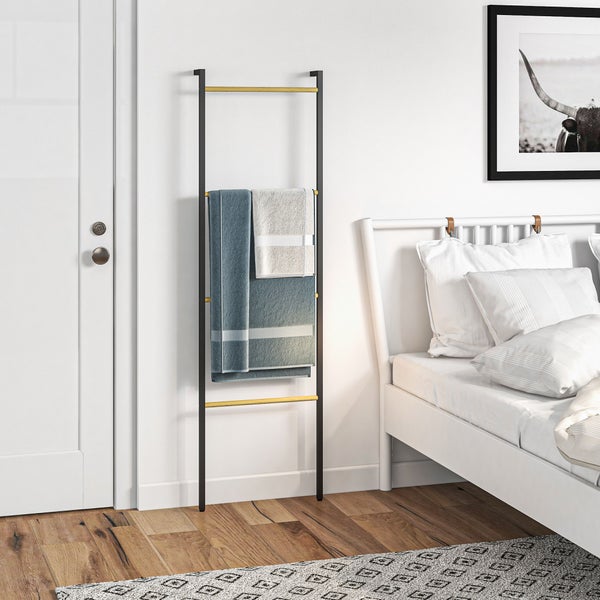 Handtuchleiter aus schwarzem Metall mit Holzsprossen lehnt in einem hellen Schlafzimmer an einer weißen Wand neben einem Bett.