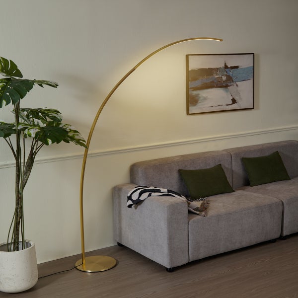 Moderne Bogenlampe aus Metall in einem Wohnzimmer mit grauem Sofa und einer Monstera-Pflanze. Die Stehlampe spendet warmes Licht.