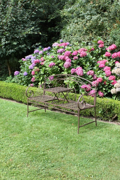 Verzierte Gartenbank aus Metall mit integriertem Tisch auf einer Rasenfläche vor blühenden Hortensien und einer niedrigen Hecke.