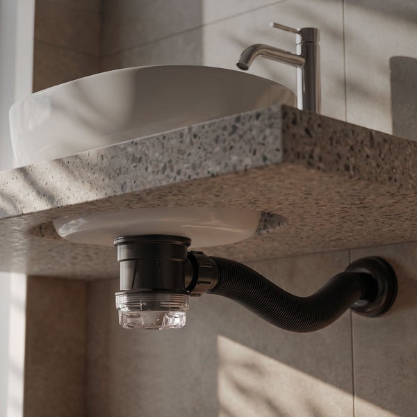 Raumsparsiphon in Schwarz mit transparentem Becher unter weißem Aufsatzwaschbecken auf einer Steinplatte im Terrazzo Design.