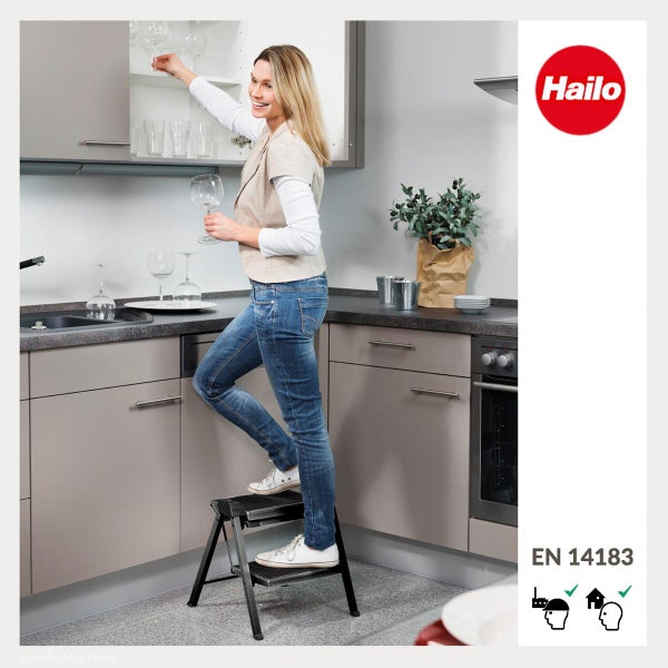 Eine Frau nutzt eine klappbare Hailo Trittleiter in einer modernen Küche, um ein Glas aus dem Schrank zu nehmen. Hailo Logo und Sicherheitsnorm.