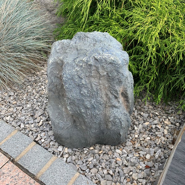 Künstlicher Gartenstein in natürlicher Felsoptik auf einem grauen Kiesbett in einem Garten mit Pflanzen.