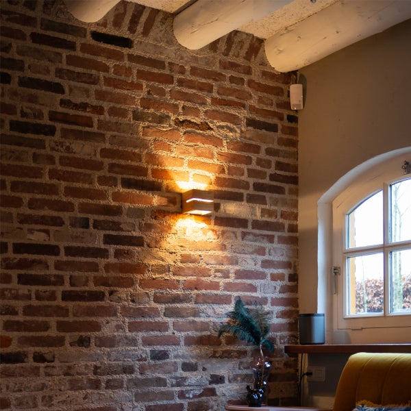 Moderne Wandleuchte aus Holz montiert an einer rustikalen Backsteinwand in einem gemütlichen Wohnraum mit Deckenbalken und Fenster.