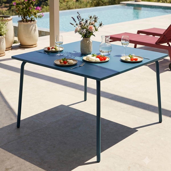 Moderner quadratischer blauer Gartentisch auf einer sonnigen Terrasse am Pool, gedeckt mit Tellern und Blumen.