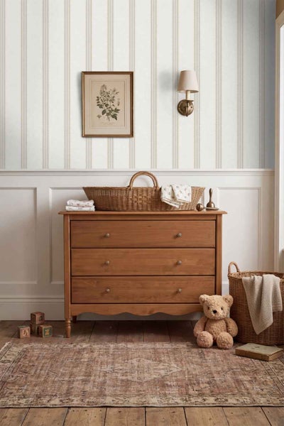 Ein Kinderzimmer mit gestreifter Tapete, einer hölzernen Kommode, einem Weidenkorb und einem Teddybären in einer klassischen, gemütlichen Umgebung.