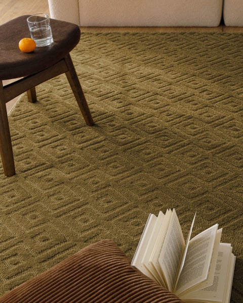 Moderner, strukturierter Teppich mit geometrischem Quadratmuster in einem Wohnzimmer mit Hocker und Buch.