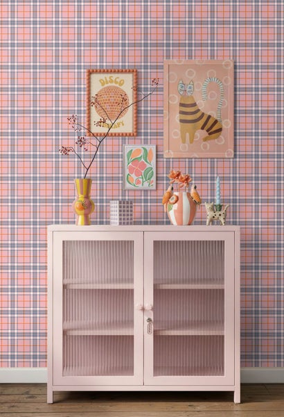 Heller Wohnraum mit karierter Tapete, einem rosa Metallschrank mit Riffelglas sowie Wandbildern und Dekorationsartikeln.