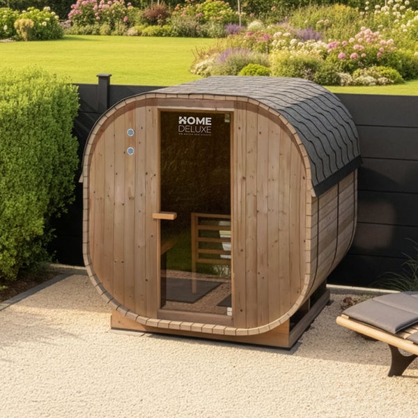 Fasssauna aus Holz von Home Deluxe Logo für den Außenbereich in einem Garten mit Kiesfläche und dunklem Sichtschutzzaun.