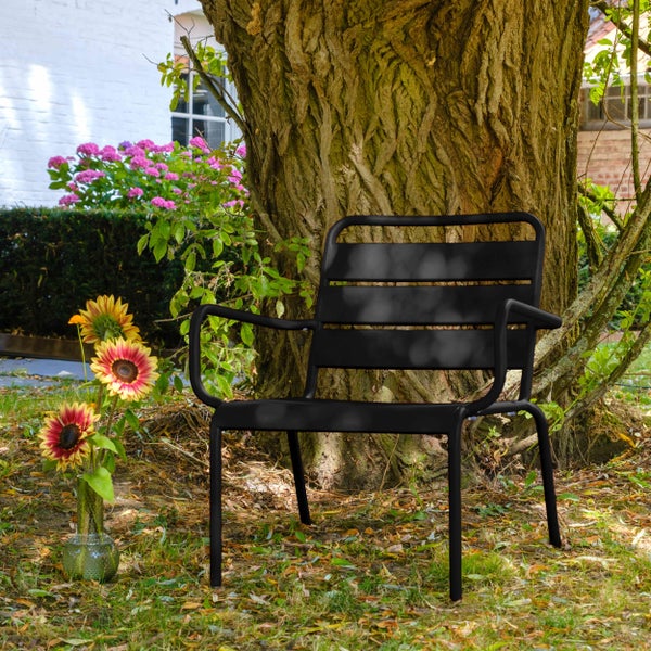 Moderner Gartenstuhl aus schwarzem Metall mit Armlehnen, platziert vor einem Baumstamm in einem hellen Garten mit Blumen.