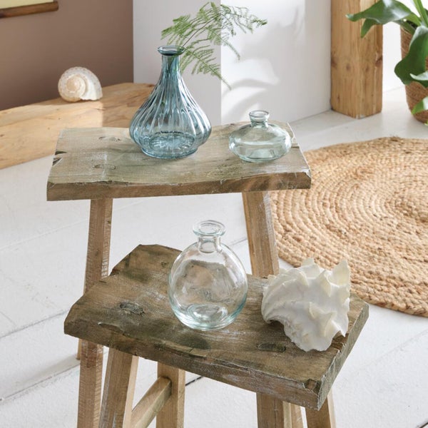 Zwei rustikale Holzhocker als Beistelltische mit verschiedenen Glasvasen und einer dekorativen Muschel in einem hellen Raum.