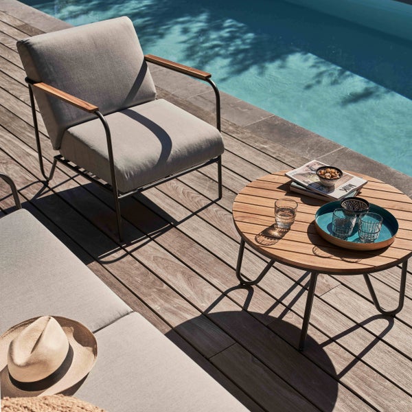 Moderne Gartenlounge mit gepolstertem Sessel und rundem Holztisch auf einer Holzterrasse am Pool.
