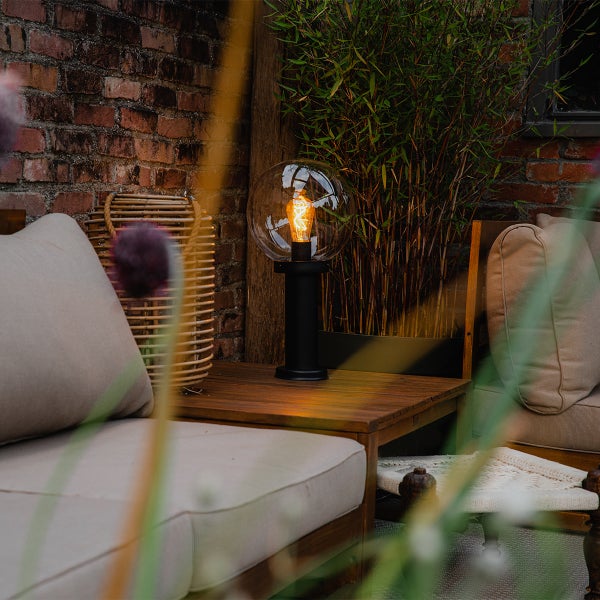 Moderne Außen-Tischleuchte mit Glaskugel auf einem Holzbeistelltisch in einer gemütlichen Gartenumgebung mit Pflanzen und Polstermöbeln.