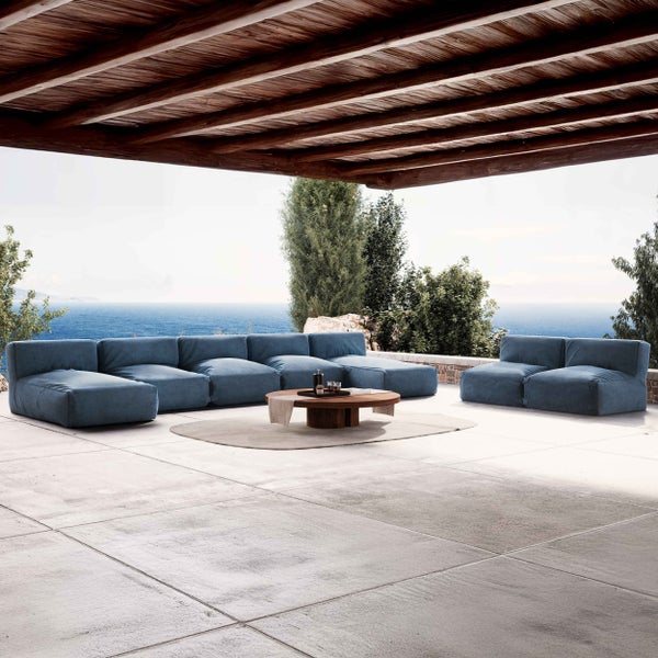 Moderne Outdoor-Lounge mit blauem Modulsofa, rundem Holztisch und Teppich auf einer hellen Steinterrasse mit Meerblick unter einem Holzdach.