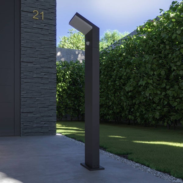 Moderne anthrazitfarbene Wegeleuchte mit Bewegungsmelder im Garten neben Rasen und Hecke, Steinwand im Hintergrund.