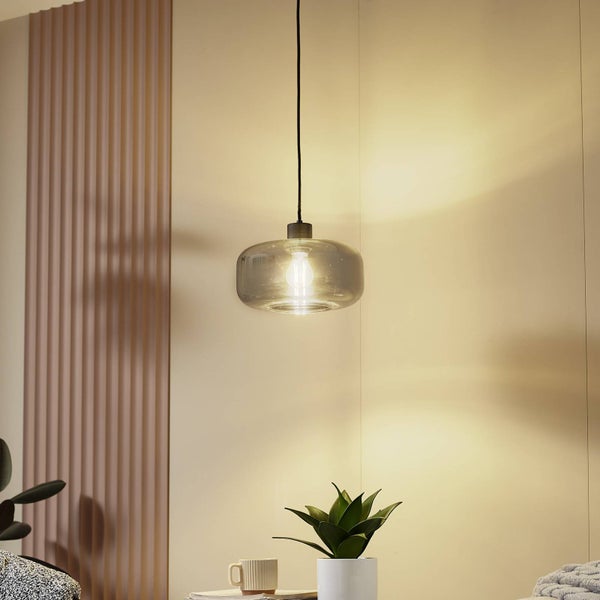 Pendelleuchte mit ovalem Schirm aus Rauchglas über einem Tisch mit Pflanze vor einer Wand mit Paneelen.