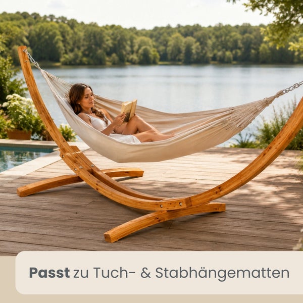 Frau liest in einer Tuchhängematte mit gebogenem Holzgestell auf einer Holzterrasse an einem See.