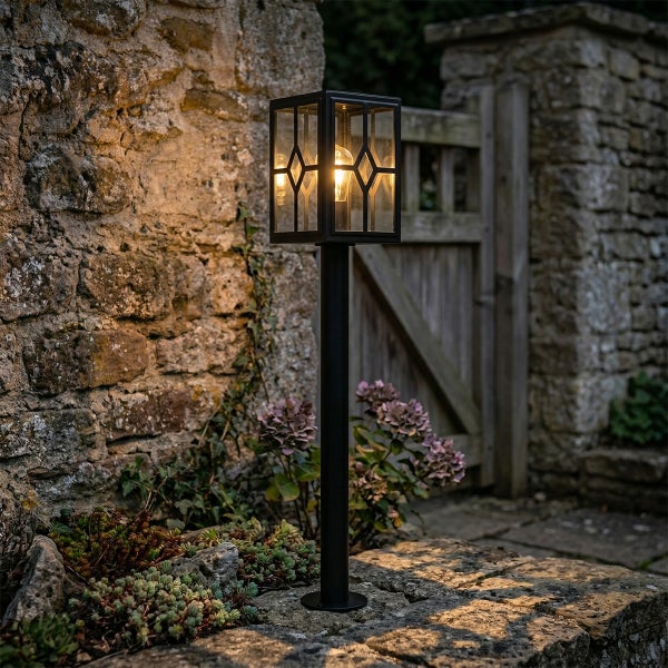 Schwarze Außenstandleuchte in Laternenform mit dekorativem Glasmuster vor einer Natursteinmauer im Garten.