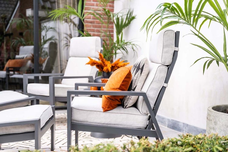 Moderne Garten-Loungestühle aus Metall mit hellgrauen Polstern und orangefarbenen Dekokissen auf einer hellen Terrasse mit Pflanzen.
