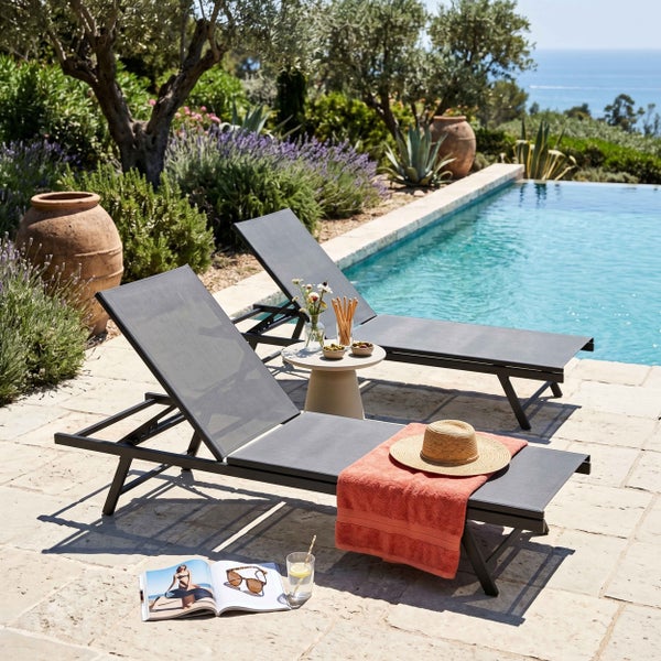 Zwei moderne graue Gartenliegen auf einer hellen Steinterrasse an einem Pool in einem mediterranen Garten.