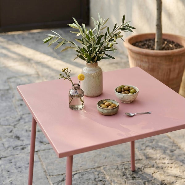 Quadratischer rosa Gartentisch aus Metall auf einer Terrasse, dekoriert mit Vasen, Olivenzweigen und Schalen mit Oliven.