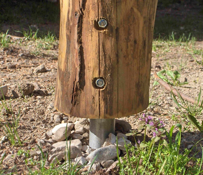 Holzpfosten mit zwei Schraubbolzen montiert auf einem Pfostenträger aus Metall im Gartenboden.