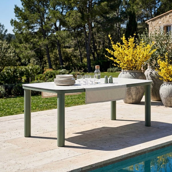 Moderner Gartentisch mit grünen Metallbeinen und heller Tischplatte auf einer Terrasse am Pool, dekoriert mit Geschirr und Gläsern.