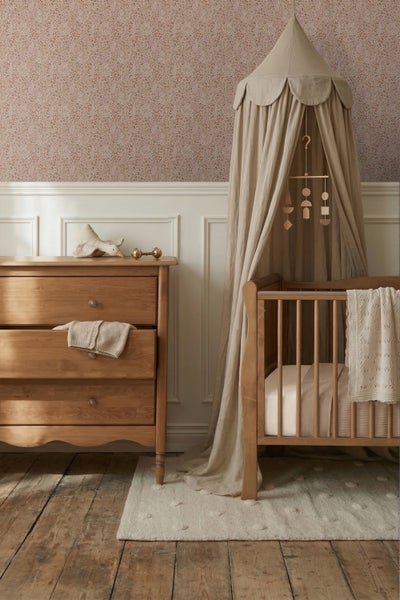 Gemütliches Babyzimmer mit Holz-Gitterbett, Stoffbaldachin, Kommode aus Holz und floraler Tapete über Wandpaneelen.