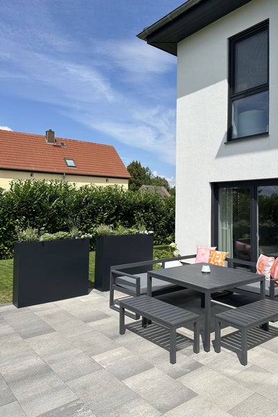 Moderne Terrasse mit dunkelgrauem Gartenmöbel Set bestehend aus Tisch, Bänken und Ecksofa sowie großen Pflanzkübeln vor einer Hecke.