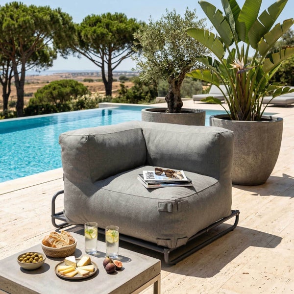 Modernes Lounge-Sofa mit Hocker auf einer Holzterrasse am Pool mit gemustertem Außenteppich und grüner Hecke im Hintergrund.